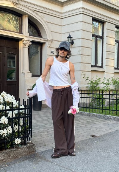 Top corto blanco, pantalones marrones con cordón, y camisa de botones a rayas. Gorro negro y gafas de sol. Sosteniendo un pequeño ramo de flores.