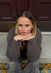 Καφέ, χοντρό πλεκτό πουλόβερ με φουσκωτά μανίκια· παντελόνι με λεοπάρ σχέδιο· μακριά, ίσια μαλλιά· στηρίζοντας τα χέρια κάτω από το πηγού�νι σε πέτρινες σκάλες.