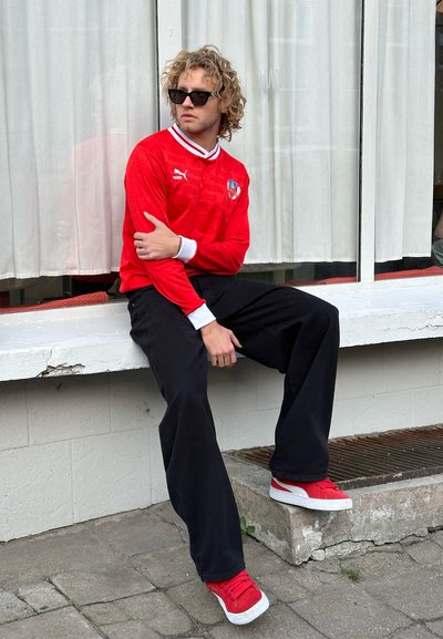 Jersey deportivo rojo de manga larga con cuello blanco y logo, combinado con pantalones sueltos negros y zapatillas rojas, sentado en un borde.