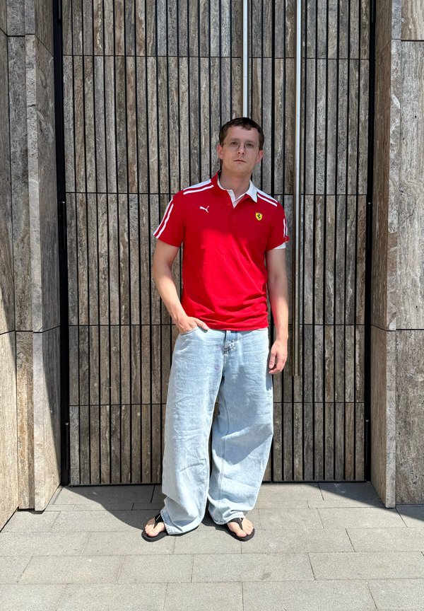 Joven con polo rojo, vaqueros claros y holgados, y sandalias, apoyado en una pared alta de listones de madera al aire libre bajo la luz del sol.