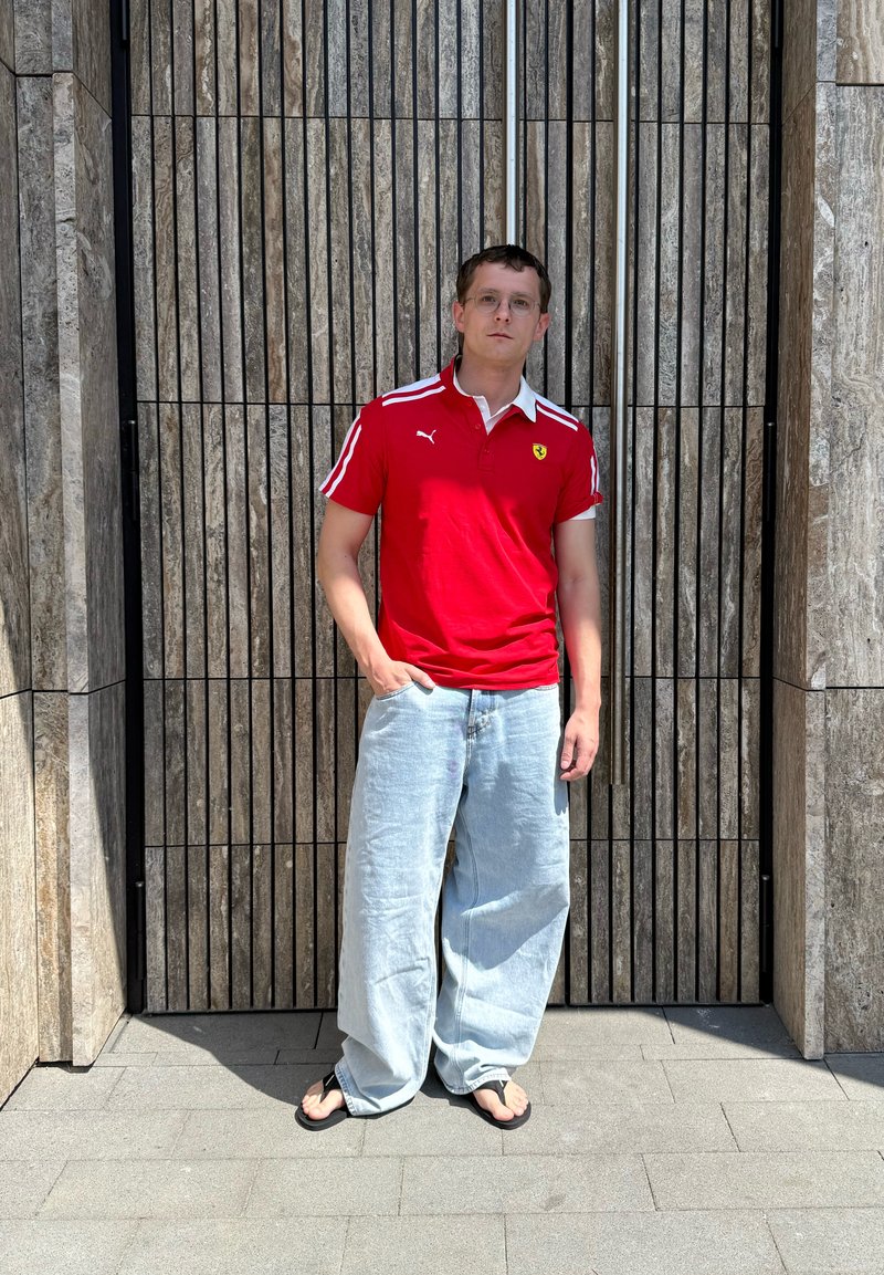Jeune homme portant un polo rouge, un jean clair ample et des tongs, debout contre un mur haut en lattes de bois à l'extérieur au soleil.