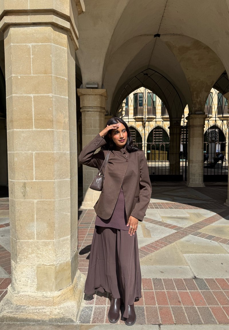 Donna con giacca marrone, gonna lunga e stivali che si tiene gli occhi ombreggiandoli in un cortile soleggiato con archi e colonne di pietra.