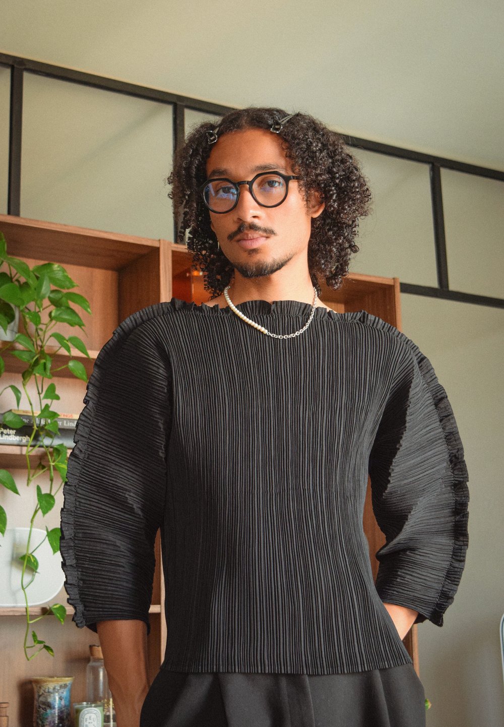Top negro con pliegues y mangas anchas, fabricado con tejido texturizado y un borde con volantes. La modelo lleva gafas y un collar sencillo, posando en un entorno neutral.