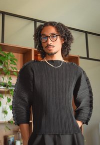 Top negro con pliegues y mangas anchas, fabricado con tejido texturizado y un borde con volantes. La modelo lleva gafas y un collar sencillo, posando en un entorno neutral.