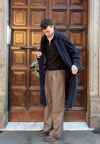 Jeune homme ajustant un long manteau sombre, portant un pantalon ample marron et un pull noir, debout devant une grande porte en bois encadrée de pierre.