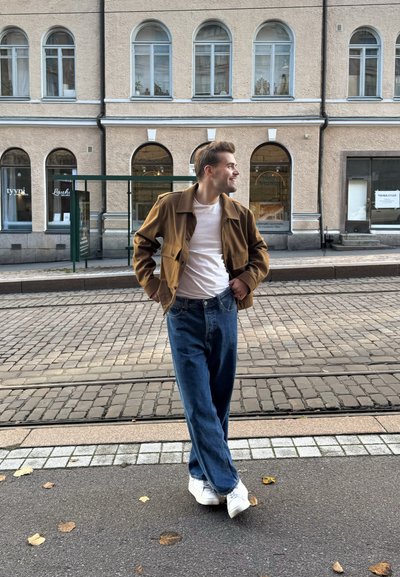 Chaqueta marrón, camiseta blanca, jeans de mezclilla azules y zapatillas blancas. De pie en una calle adoquinada cerca de edificios con ventanas arqueadas.