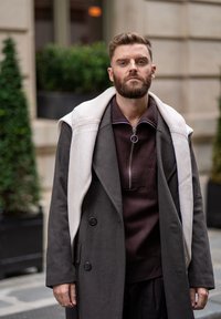 Szary wełniany płaszcz z dwoma guzikami, nałożony na ciemnobrązowy sweter dzianinowy z zamkiem przy kołnierzu, oraz jasnobeżowy szalik swobodnie zarzucony na ramionach.