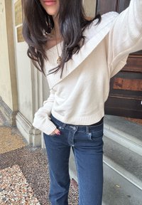 Pull crème, décolleté bretelles, avec des poignets côtelés, associé à un jean foncé ajusté avec un léger évasement et des poches standard.