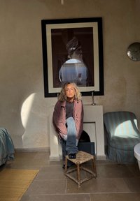 En person med krøllet hår sidder på en flettet skammel iført en ternet overshirt og jeans. Et indrammet portræt hænger på en struktureret væg.