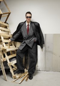 Veste de costume croisée grise portée sur une chemise blanche et une cravate rouge, associée à un pantalon en jean noir surdimensionné et des chaussures en cuir noires. Lunettes de soleil noires.