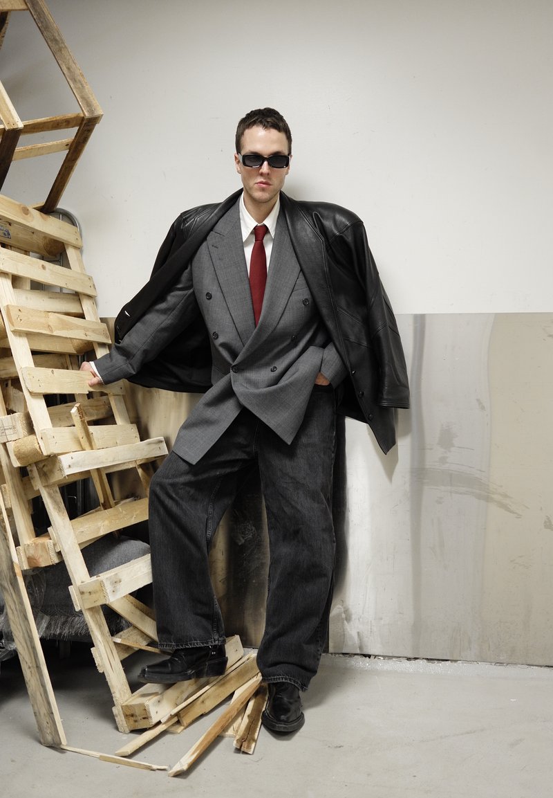 Veste de costume croisée grise portée sur une chemise blanche et une cravate rouge, associée à un pantalon en jean noir surdimensionné et des chaussures en cuir noires. Lunettes de soleil noires.