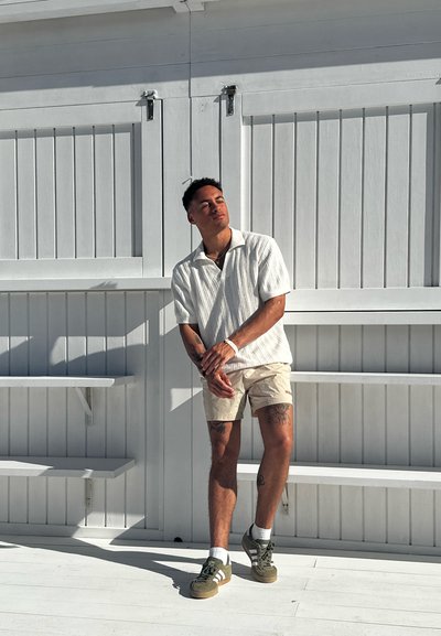 Camisa de rayas blanca de manga corta, pantalones cortos beige, zapatillas verdes, de pie contra un fondo de madera blanca con paneles horizontales.