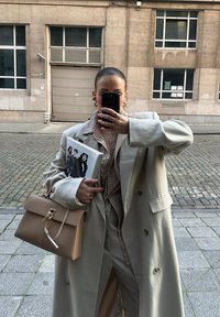 Beige dubbele-breasted jas over een geruit overhemd, met een tijdschrift en een beige handtas met een gouden sluiting. Gladde, elegante texturen.