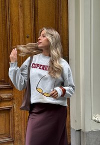 Szary sweter z napisem "COPENHAGEN" w bordo, w zestawie z bordową spódnicą. Trzyma żółte okulary przeciwsłoneczne. Tło w drewnianych panelach.