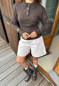 Persona che indossa un maglione marrone a maniche lunghe con bottoni, pantaloni corti bianchi, stivali neri e gioielli d'oro, in piedi su una terrazza di legno vicino a una porta.