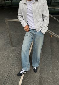 Camisa de botão listrada em cinza claro e branco, por cima de uma t-shirt branca, combinada com jeans largos em azul claro e ténis pretos com detalhes brancos.