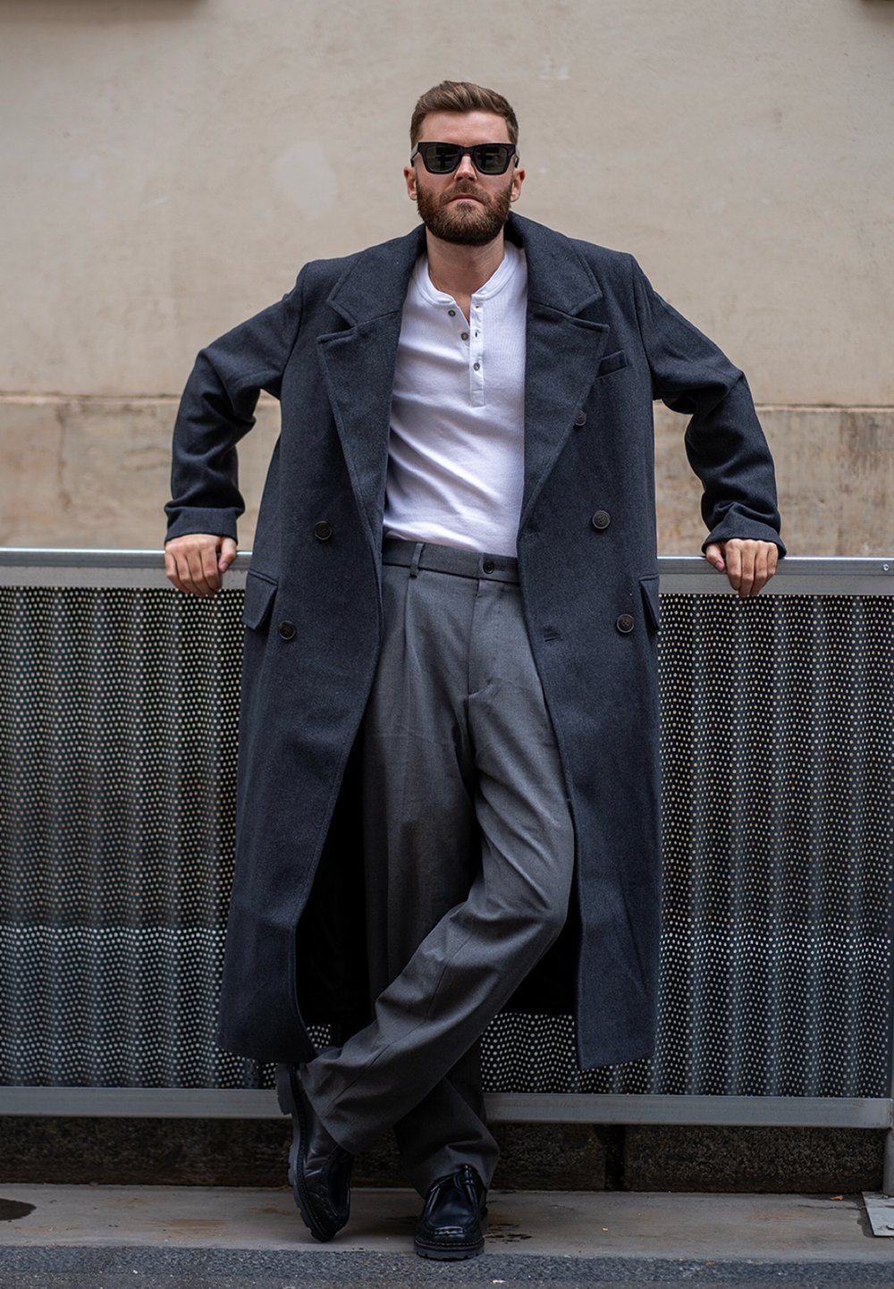 Uomo che indossa un soprabito di lana grigio scuro, una maglia henley bianca, pantaloni grigi e scarpe nere. Appoggiato a una superficie di rete metallica.