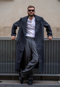 Uomo che indossa un soprabito di lana grigio scuro, una maglia henley bianca, pantaloni grigi e scarpe nere. Appoggiato a una superficie di rete metallica.