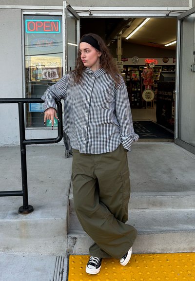 Persona joven apoyada en una barandilla fuera de la entrada abierta de una tienda, vistiendo una camisa a cuadros, pantalones anchos y zapatillas deportivas.