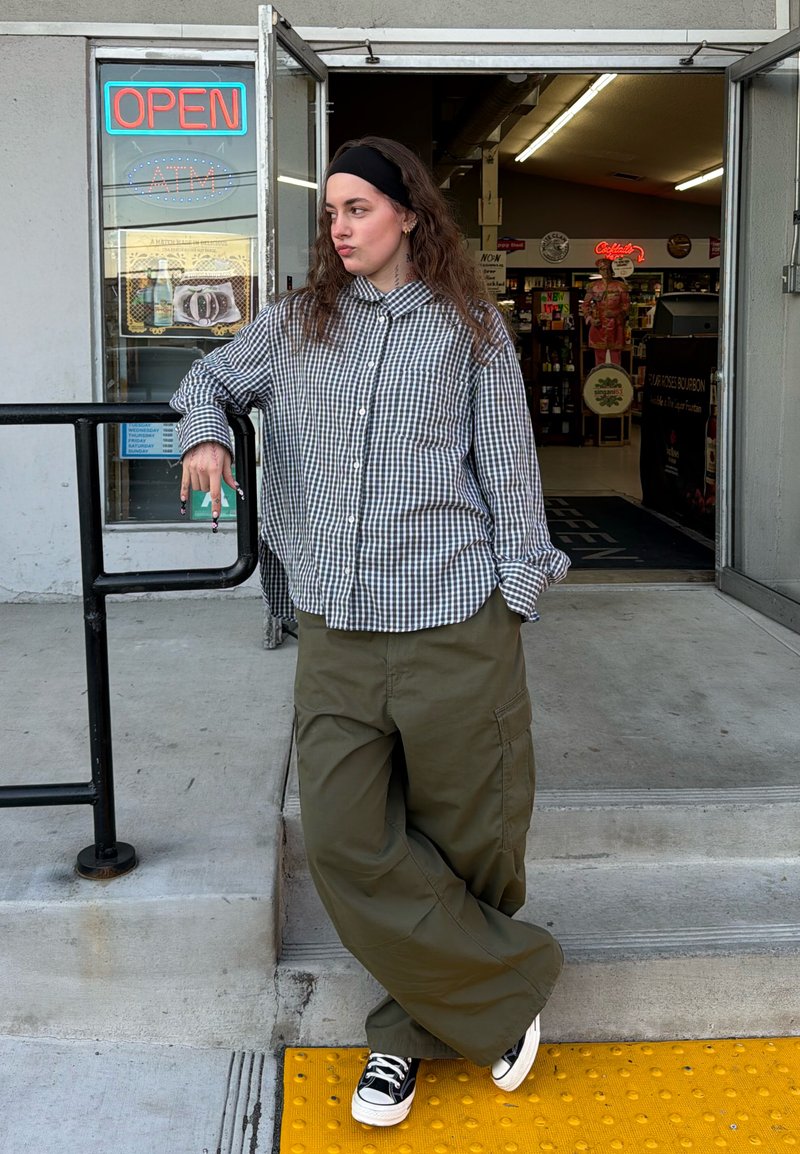 Jeune personne appuyée sur une rampe à l'extérieur d'une entrée de magasin ouverte, portant une chemise à carreaux, un pantalon large et des baskets.