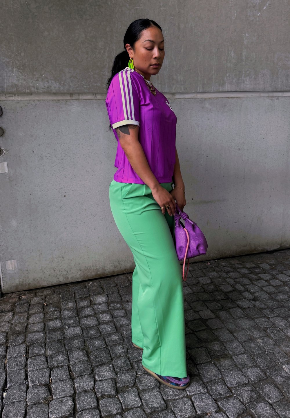 Jersey viola adidas con strisce bianche, abbinato a pantaloni svasati verdi. Tiene una piccola borsa viola con texture. Capelli neri, sfondo neutro.