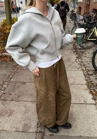 Hellgrauer Kapuzenpullover mit Reißverschluss, oversize Passform, kombiniert mit lockeren, zerknitterten braunen Hosen. Die Person hält einen grünen Kaffeebecher.