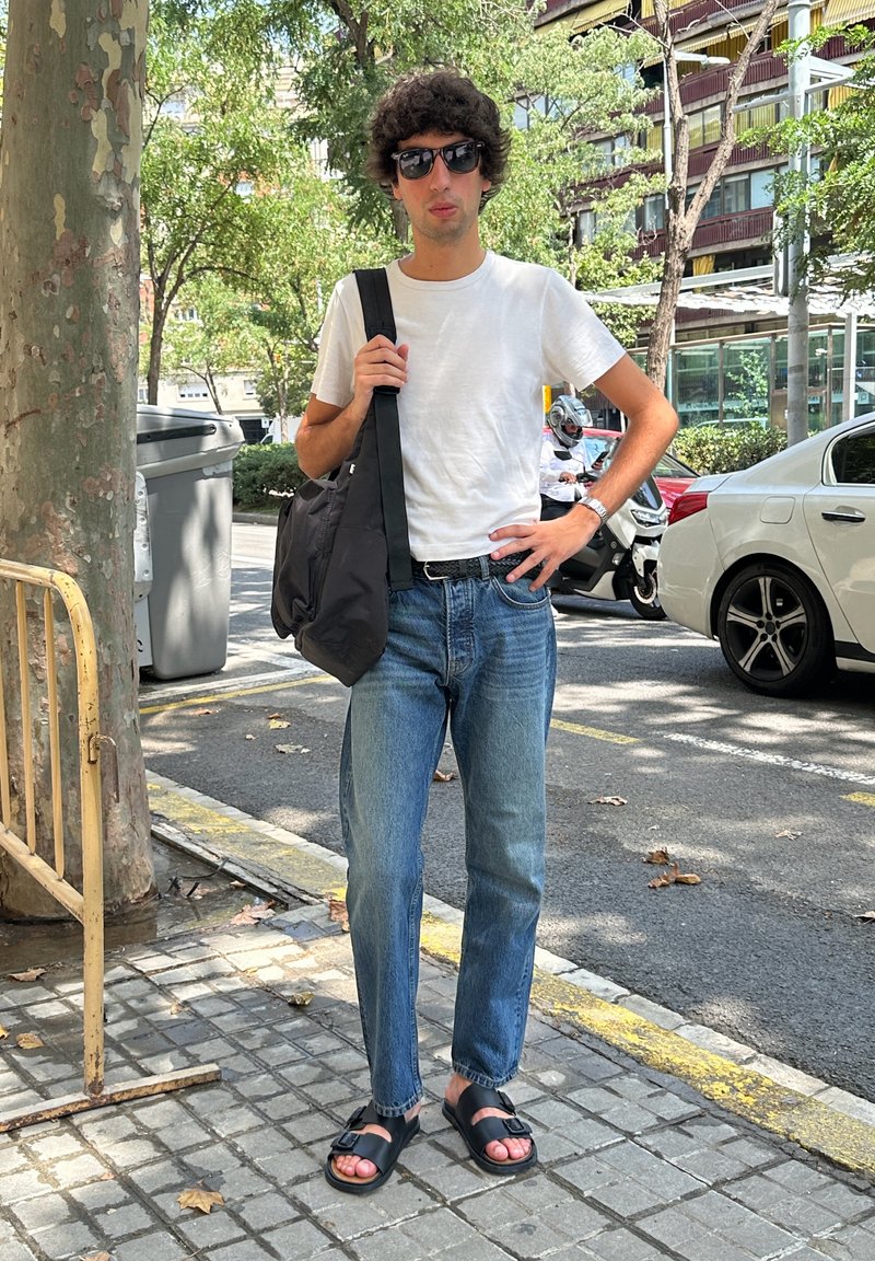 Junger Mann mit Sonnenbrille, weißem T-Shirt, Jeans und schwarzen Sandalen steht auf einem städtischen Gehsteig und hält eine schwarze Schultertasche.