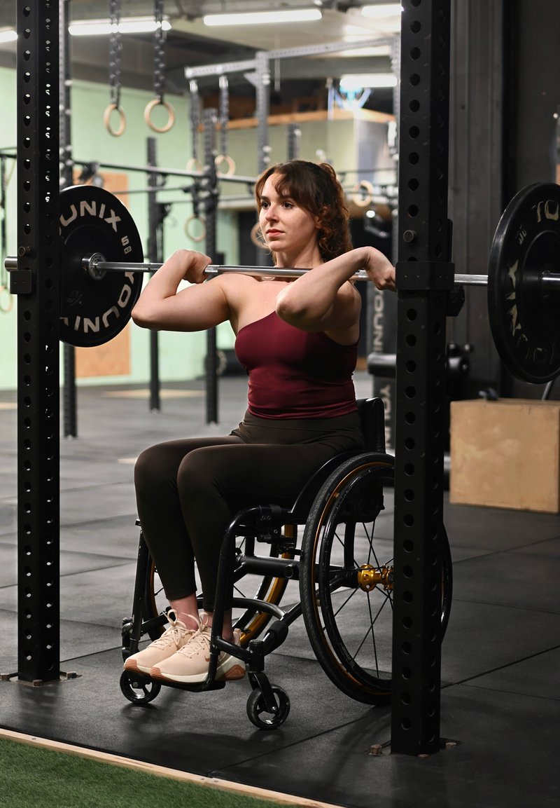 Un utilisateur de fauteuil roulant effectue un exercice avec une barre, portant un haut bordeaux et des leggings foncés, dans une salle de sport avec un sol en caoutchouc noir et du matériel d'haltérophilie.