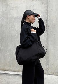 Bolso de tela negro y oversize con una textura suave. Llevado con una blusa negra a juego con un alto cuello. Incluye una gorra negra.