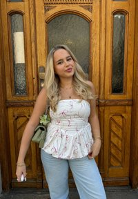 Trägerloses weißes Oberteil mit rosa Bandmuster, ausgestellter Saum; hellblaue Jeans; grüne Handtasche mit floralen Details; steht gegen eine Holztür.