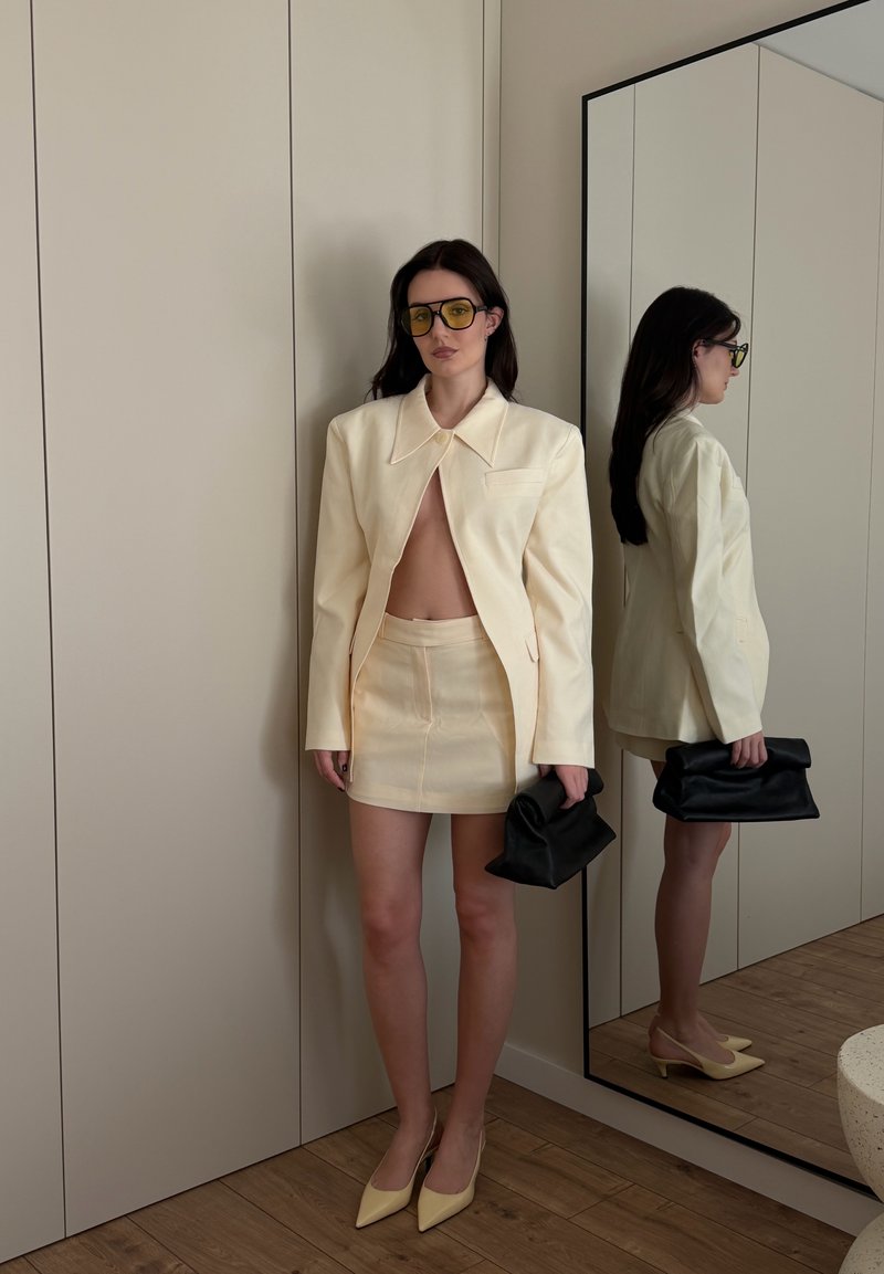 Donna che indossa blazer e gonna giallo pallido, occhiali con lenti gialle, che tiene in mano una pochette nera, in piedi accanto a uno specchio a figura intera in una stanza neutra.