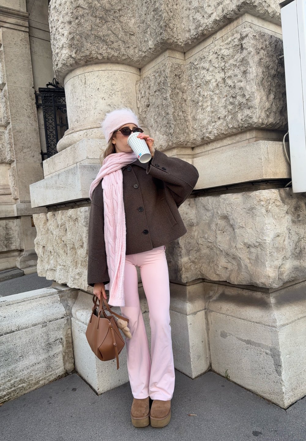 Hnědý vlněný kabát s velkými knoflíky, růžový pletený šátek, růžové křivákové kalhoty, hnědá kabelka a platformové boty. Stojící proti texturovanému kameni.