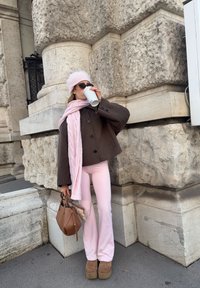 Hnědý vlněný kabát s velkými knoflíky, růžový pletený šátek, růžové křivákové kalhoty, hnědá kabelka a platformové boty. Stojící proti texturovanému kameni.