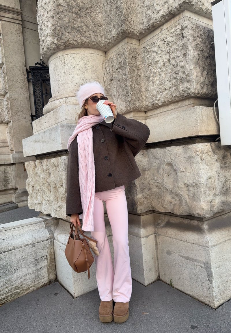 Brauner Wollmantel mit großen Knöpfen, rosa Strickschal, rosa ausgestellte Hose, braune Handtasche und Plateauschuhe. Stehend vor strukturiertem Stein.