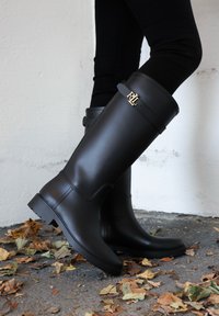 Melni gumijas ceļgalu zābaki ar gludu virsmu, rotāti ar zelta logo uz siksniņas un ar teksturētu zolīti, novietoti uz lapām noklātas virsmas.