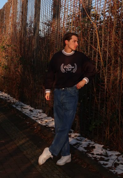 Sudadera negra con letras blancas y cuello acanalado, combinada con jeans azules holgados y zapatillas blancas. El fondo tiene vegetación y nieve.