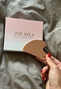 ZOE AYLA gua sha įrankis rožių auksu, kreivai formos su lygiu paviršiumi, laikomas šalia rožinės dėžutės su užrašu "ZOE AYLA® IŠSKIRTINĖ KOLEKCIJA x ZALANDO."