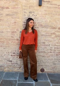 Rostfarbener V-Ausschnitt-Pullover mit strukturierten Details, kombiniert mit weit geschnittenen, braunen Cordhosen. Hält eine kleine braune geflochtene Tasche. Steinmauer im Hintergrund.