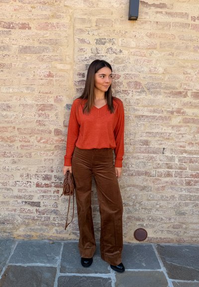 Suéter naranja de pico con detalles texturizados, combinado con pantalones de pana marrón de pierna ancha. Sosteniendo un pequeño bolso de mimbre marrón. Fondo de pared de piedra.