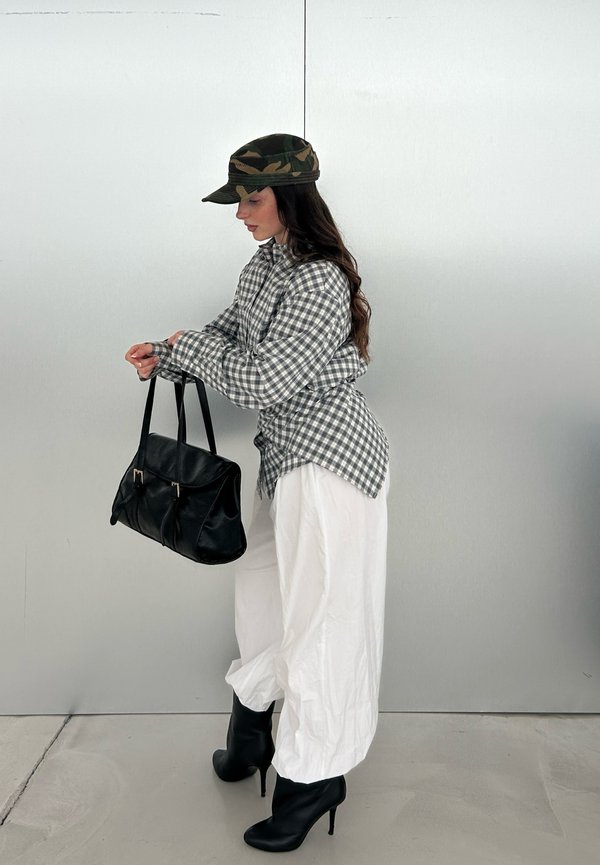 Femme en bottes à talons noires, pantalon blanc ample, chemise grise à carreaux, casquette camouflage, tenant un sac à main noir contre un mur clair uni.