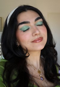 Kvinne med langt svart hår som har på seg en grønn topp. Utpregede trekk inkluderer mynteøyenskygge, blanke lepper, og lagdelt gull- og lilla smykker.