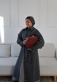Femme portant un foulard noir à motifs et un manteau gris à carreaux, souriant, tenant un sac à main marron texturé, debout devant un canapé gris clair.