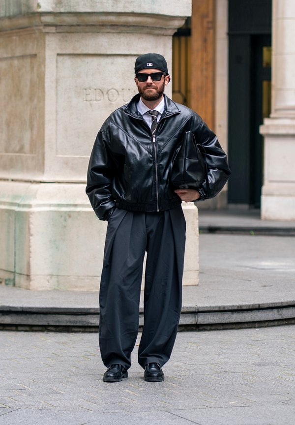 Man wearing black leather jacket, black wide trousers, black cap, sunglasses, and holding a black leather bag, standing on a city street.