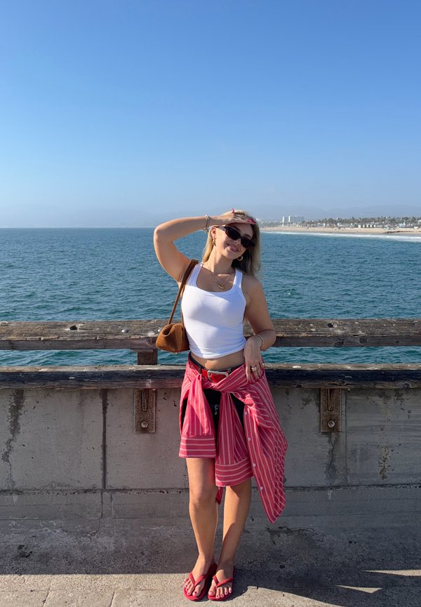 Una mujer con gafas de sol posa junto a una barandilla de madera en un muelle junto al mar y se protege los ojos del sol, mientras el océano y la costa se extienden a su espalda.