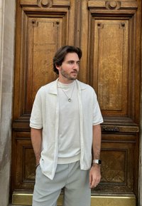 Camisa blanca de manga corta y botones con un patrón texturizado sobre una camiseta blanca de cuello redondo, combinada con pantalones claros de rayas finas grises.