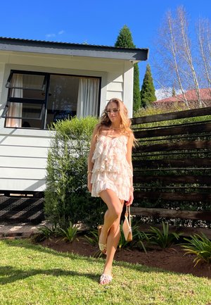 Vestido beige claro en capas con volantes, combinado con sandalias. El fondo presenta una casa blanca, arbustos verdes y una cerca de madera. Día soleado y brillante.