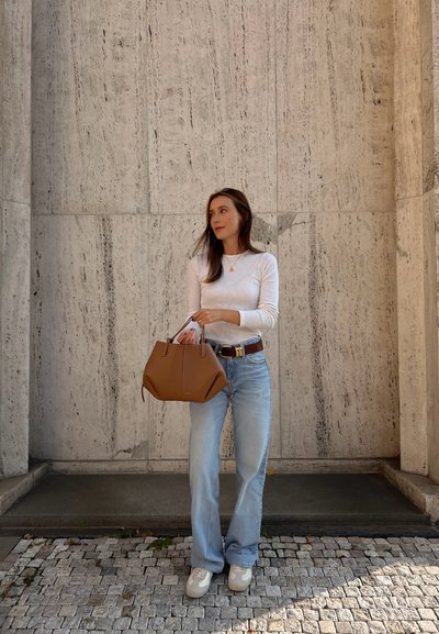 Bolso de cuero marrón con diseño estructurado y asas dobles, combinado con jeans acampanados de color azul claro y una blusa blanca de manga larga.