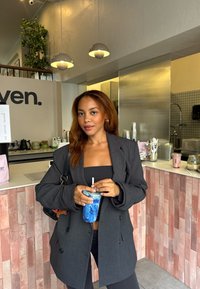 Kullgrå, dobbeltspent blazer over en svart topp, holder en blå drink med sugerør, mot en lys kafébakgrunn.