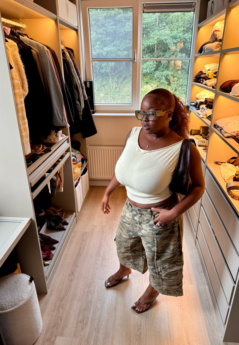 Femme portant un haut blanc et un short camouflage, debout dans un dressing avec des vêtements accrochés, des étagères et une grande fenêtre donnant sur de la verdure extérieure.