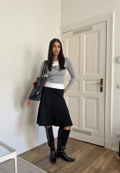 Mujer con cabello largo y oscuro que lleva una blusa gris y blanca, falda negra, botas negras hasta la rodilla y un bolso negro grande, en el interior junto a una puerta cerrada.